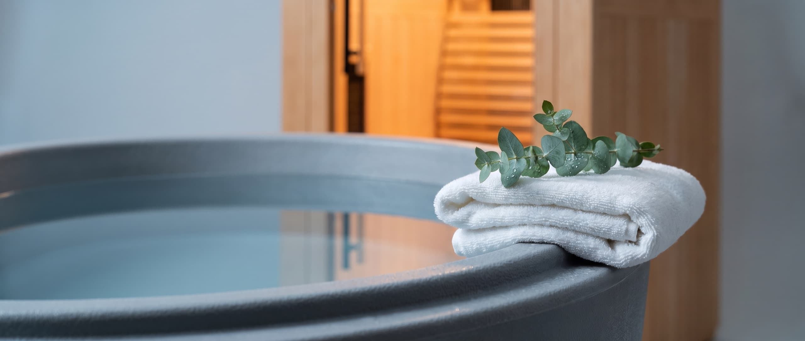 Le rebord d'un bain froid à l'eau immobile, une serviette blanche et un brin d'eucalyptus, devant la porte vitrée d'un sauna éclairé
